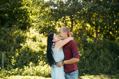 A couple is hugging in a lush green forest. The man is wearing a red plaid shirt