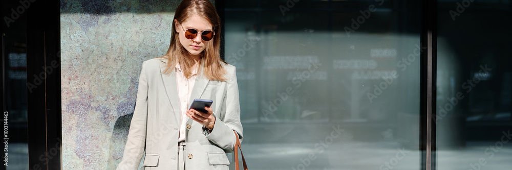 Header image of young female manager wearing fashionable outfit using smartphone while standing outdoors against office building in business district Header image of young female manager wearing fashionable outfit using smartphone while standing outdoors against office building in business district