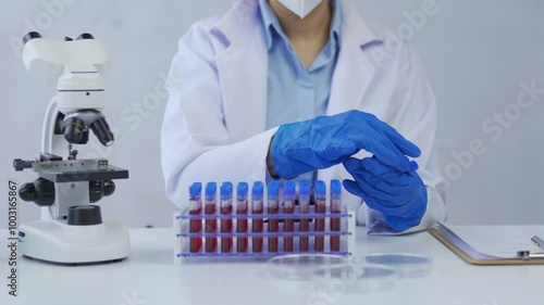 Scientist in a lab coat and blue gloves picks up a blood sample tube from a rack in a laboratory setting