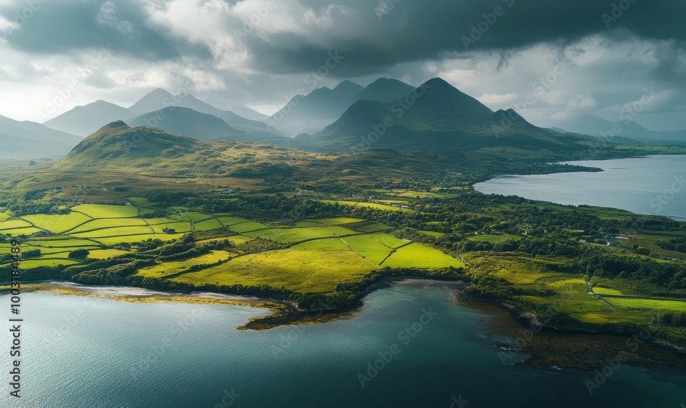 Obraz premium Aerial view of tranquil shoreline, green fields, and majestic mountains under a cloudy sky
