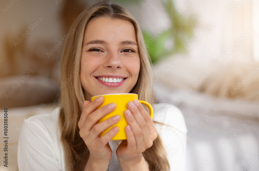 A young woman smiles brightly while holding a yellow mug close to her face. She is in a cozy, warmly lit bedroom with soft bedding and greenery in the background, evoking happiness and relaxation. A young woman smiles brightly while holding a yellow mug close to her face. She is in a cozy, warmly lit bedroom with soft bedding and greenery in the background, evoking happiness and relaxation.