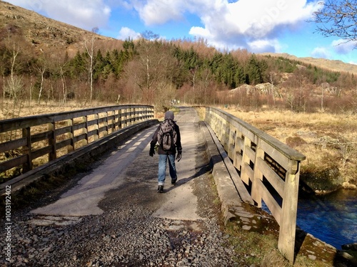 A lone boy hiker with a backpack walking across a wooden bridge in a mountainous area, capturing a sense of adventure and the journey through nature’s wild terrain. photo