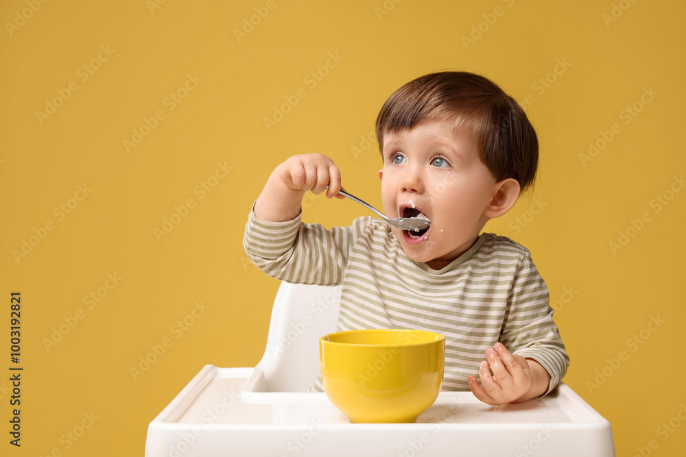Cute little kid eating healthy baby food from bowl in high chair on yellow background Cute little kid eating healthy baby food from bowl in high chair on yellow background