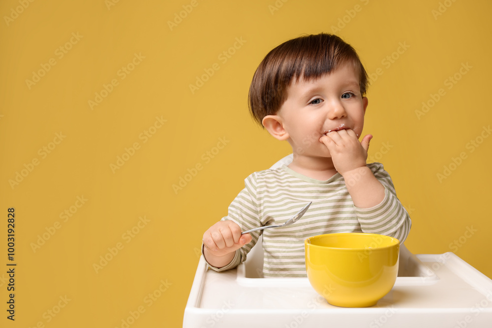 Cute little kid eating healthy baby food from bowl in high chair on yellow background, space for text Cute little kid eating healthy baby food from bowl in high chair on yellow background, space for text