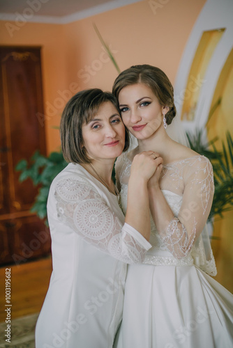 A bride is standing in front of a wedding dress, touching it and adjusting it. The dress is white and has a train. The bride is wearing a white robe and a tiara