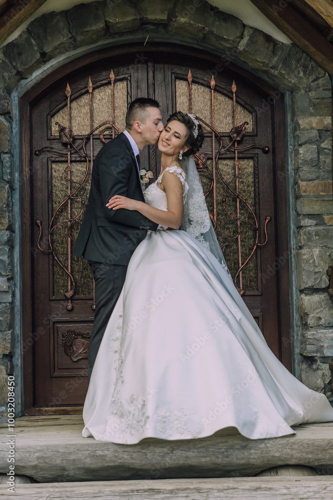 A bride and groom are kissing each other on the cheek in front of a wooden door. The bride is wearing a white dress and the groom is wearing a suit. Scene is romantic and happy A bride and groom are kissing each other on the cheek in front of a wooden door. The bride is wearing a white dress and the groom is wearing a suit. Scene is romantic and happy