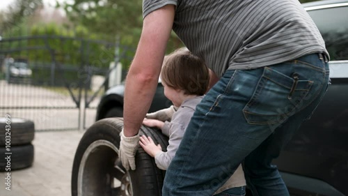 Cute toddler boy helping his father to change car wheels in their backyard. Father teaching his little son to use tools. Active parent of a small child. Slow motion summer day footage.