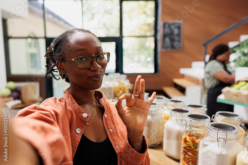 Black woman wearing spectacles doing a video advertisement of local eco friendly shop. African american female customer showing ok sign gesture in her vlog at bio food convenience store. photo