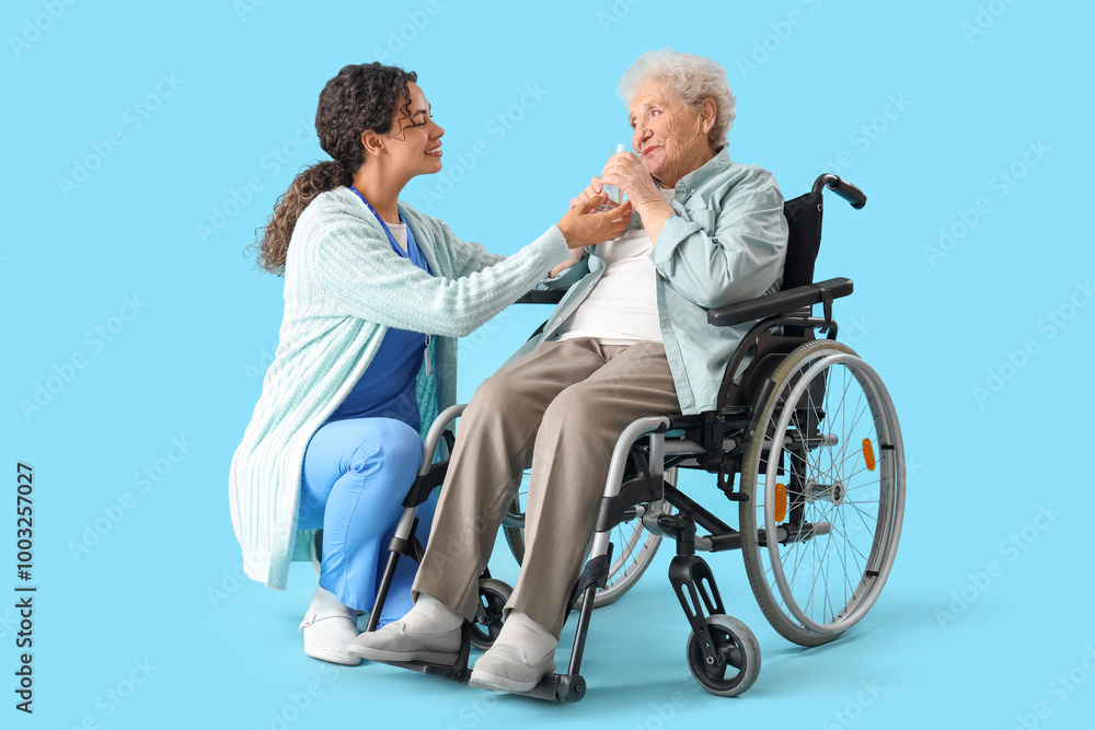 Young African-American female medical worker giving glass of water elderly woman on wheelchair against blue background Young African-American female medical worker giving glass of water elderly woman on wheelchair against blue background