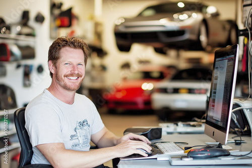 Car mechanic, smiling, working on computer in modern garage, surrounded by cars, tools, equipment, professional environment concept