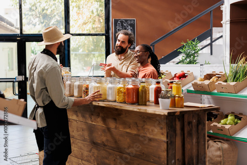 Male vendor offering advice to clients who are shopping for sustainable merchandise at eco friendly store. Owner of local supermarket describing food items in jars to the young multiracial couple.
