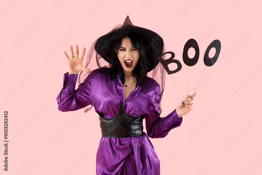 Beautiful young scary woman dressed as witch for Halloween with word BOO on pink background Beautiful young scary woman dressed as witch for Halloween with word BOO on pink background