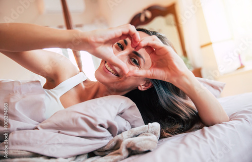 Canvas Print Lovely woman lying in bed, smiling and making a heart shape with her hands in a bright, cozy bedroom
