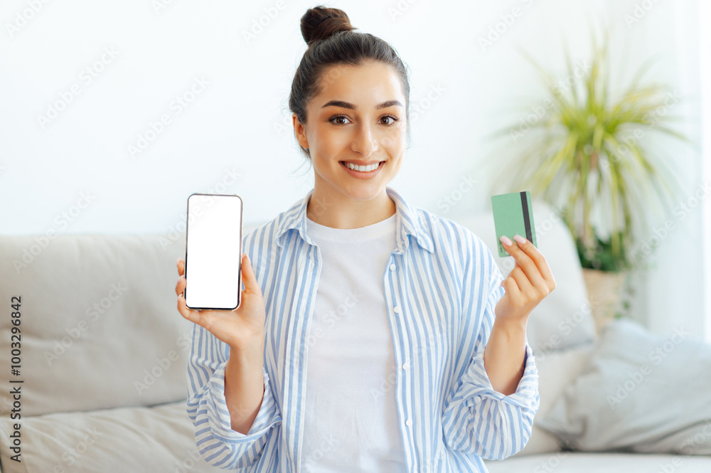 Positive attractive brunette woman, sits on a sofa in a living room, holds smartphone and credit card, shows white empty mock-up phone screen for advertising or announcement, smiles at camera Positive attractive brunette woman, sits on a sofa in a living room, holds smartphone and credit card, shows white empty mock-up phone screen for advertising or announcement, smiles at camera