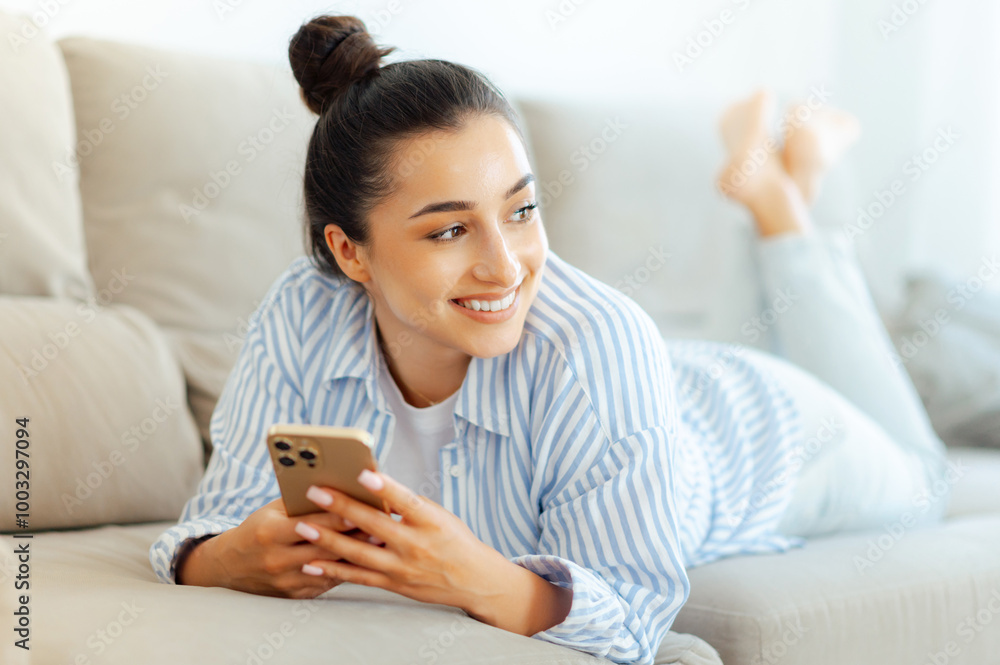 Happy young indian or arabian pretty woman relaxing at home, she is lying down on sofa, using mobile smartphone, messaging online with friends, doing online shopping, scrolling news, looks away, smile Happy young indian or arabian pretty woman relaxing at home, she is lying down on sofa, using mobile smartphone, messaging online with friends, doing online shopping, scrolling news, looks away, smile