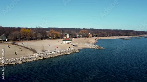 Aerial orbit shot Lake Ontario's shore. Scenery of a little cabin and people walking on the sand while enjoying the view. Beautiful sunny day and a bright blue sky with a forest in the background.