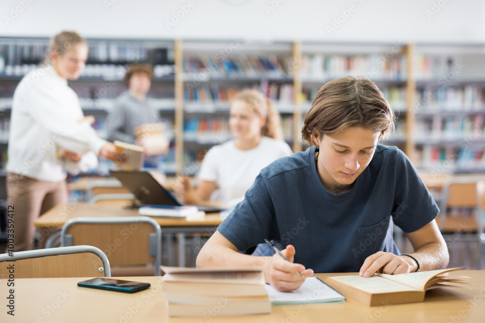 Fototapeta premium Teenage boy reading books and doing homework in library.