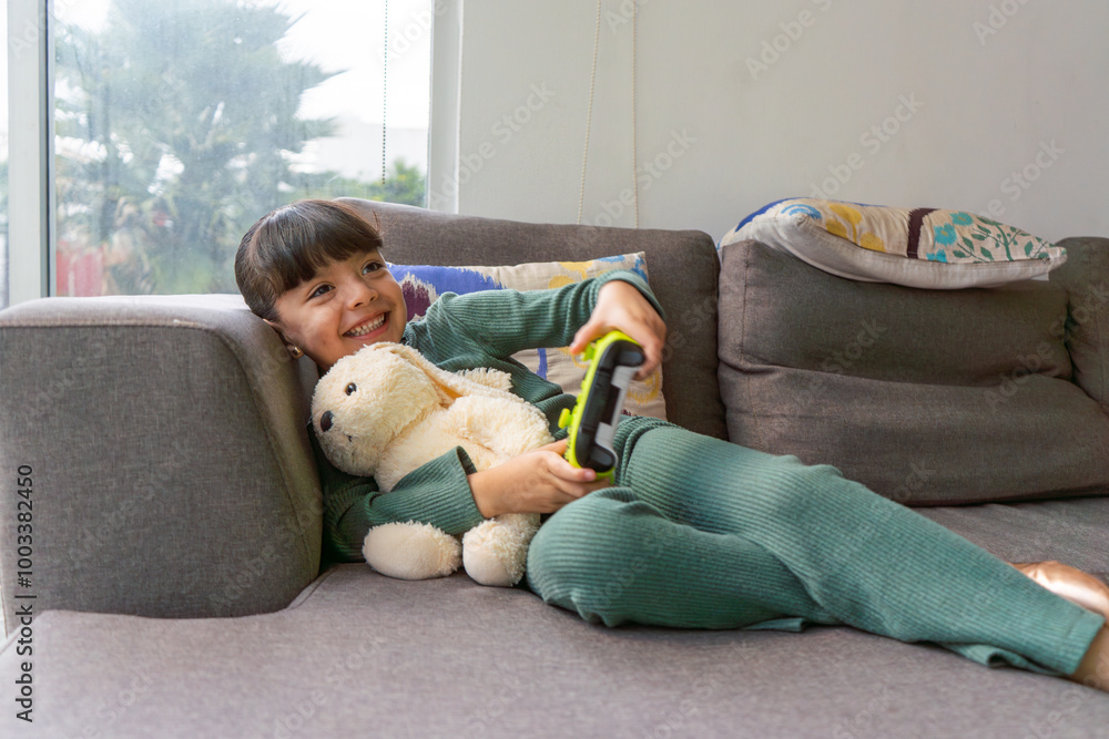 Obraz premium little girl playing video games lying on the sofa, happy kid smiling enjoying entertainment with technology