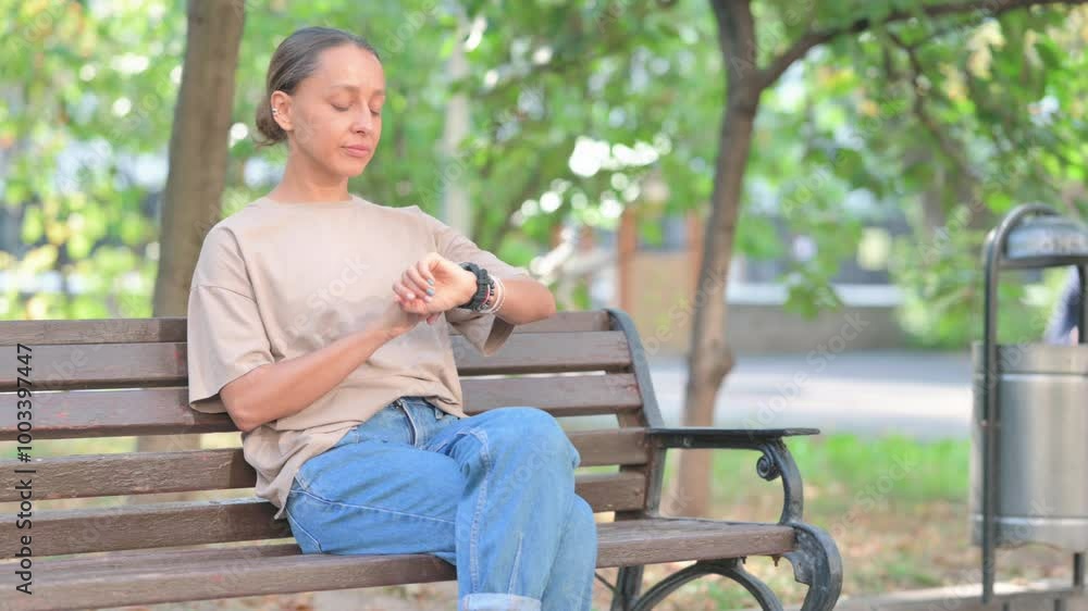 Young Hispanic Woman Using Smart Watch while Sitting Outdoor on Bench Young Hispanic Woman Using Smart Watch while Sitting Outdoor on Bench