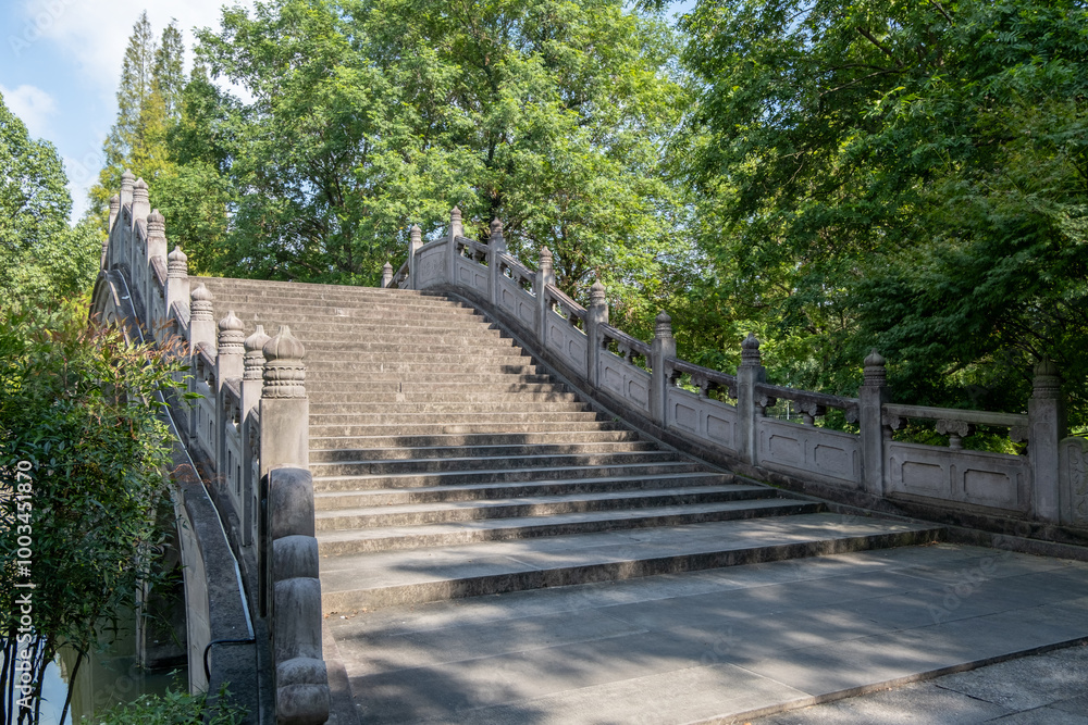 A traditional Chinese curved stone bridge with a staircase, intricate carvings, and railings in a public park or garden. Footpath and recreational infrastructure in China. A traditional Chinese curved stone bridge with a staircase, intricate carvings, and railings in a public park or garden. Footpath and recreational infrastructure in China.