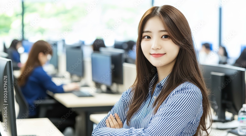 A young woman in a blue striped shirt smiles confidently in an office, embodying professionalism amidst team collaboration. A young woman in a blue striped shirt smiles confidently in an office, embodying professionalism amidst team collaboration.