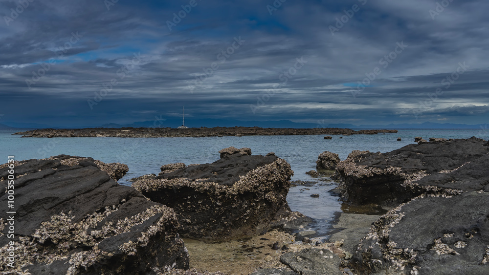Littoral at low tide. Many attached clam shells can be seen on the exposed picturesque boulders. The yacht is far away, in the turquoise ocean. A tall mast against the sky and clouds. Madagascar. Littoral at low tide. Many attached clam shells can be seen on the exposed picturesque boulders. The yacht is far away, in the turquoise ocean. A tall mast against the sky and clouds. Madagascar.