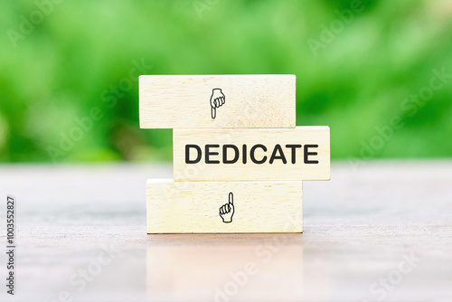 DEDICATE word written on wooden bars on a green background of plants out of focus