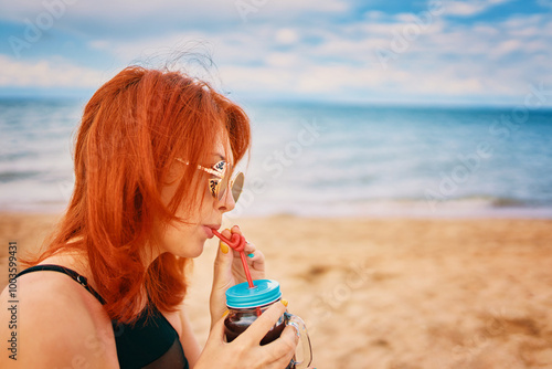 Red-haired woman with sunglasses on vacation. Holidays at the sea. Calm lady in a swimsuit is sitting on beach with a cocktail and look away. Tropical alcohol beverage. Summer day with cloudy sky.