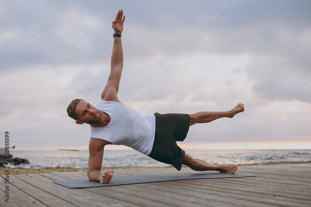 Young strong sporty athletic toned fit sportsman man wear sports clothes do side plank exercise pilates warm up train at sunrise sun dawn over sea beach outdoor on pier seaside in summer day morning. Young strong sporty athletic toned fit sportsman man wear sports clothes do side plank exercise pilates warm up train at sunrise sun dawn over sea beach outdoor on pier seaside in summer day morning.