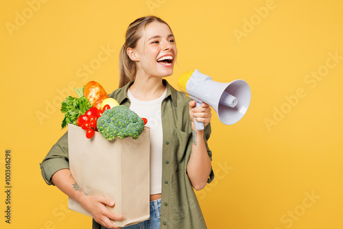 Young woman wears casual clothes hold bag for takeaway mock up with food products scream in megaphone announces discounts sale Hurry up isolated on plain yellow background. Delivery service from shop. photo