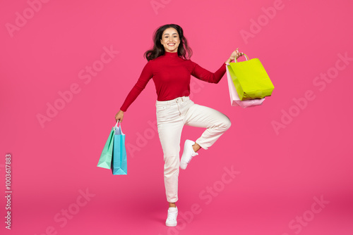 A cheerful woman joyfully jumps while holding colorful shopping bags. She wears a stylish red top and white jeans, showcasing a vibrant expression against a bright pink background.