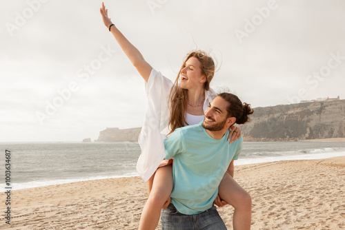 A couple shares a playful moment at the beach, with one partner giving a piggyback ride while laughing and waving. The ocean waves and soft sand create a lively, joyful atmosphere.