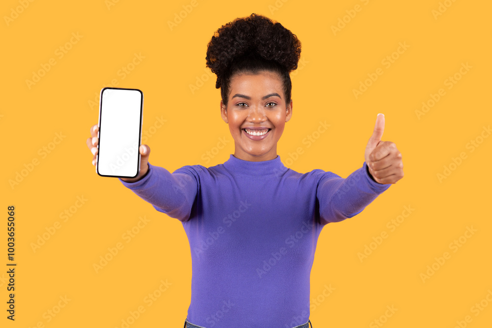 A cheerful young woman stands against a vibrant yellow backdrop, proudly holding up her smartphone with blank screen while giving a thumbs-up. Her happy expression conveys excitement about the device. A cheerful young woman stands against a vibrant yellow backdrop, proudly holding up her smartphone with blank screen while giving a thumbs-up. Her happy expression conveys excitement about the device.