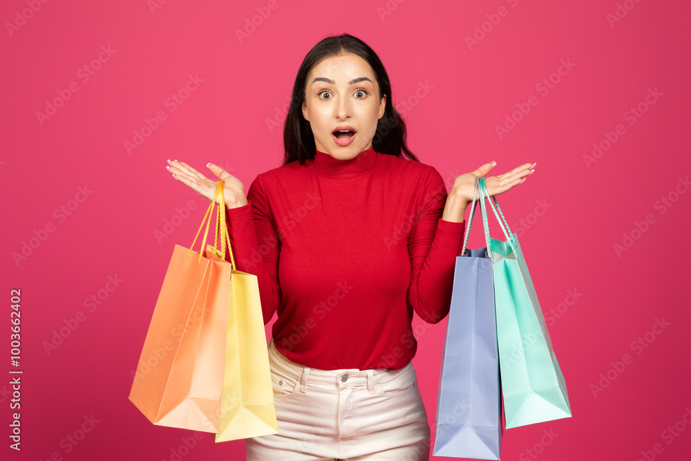 A woman in a red top displays her joy while holding two shopping bags in different colors. Behind her is a bright pink background, adding to the cheerful atmosphere of her shopping experience. A woman in a red top displays her joy while holding two shopping bags in different colors. Behind her is a bright pink background, adding to the cheerful atmosphere of her shopping experience.