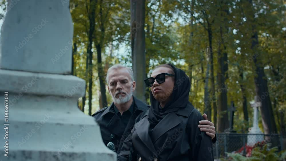 Tilt down shot of family couple visiting grave in cemetery. Man embracing his grieving wife dressed black and wearing sunglasses as looking thoughtfully at stone cross Tilt down shot of family couple visiting grave in cemetery. Man embracing his grieving wife dressed black and wearing sunglasses as looking thoughtfully at stone cross