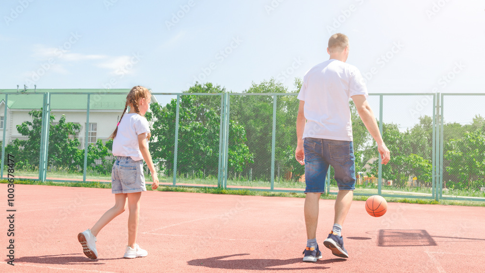 Obraz premium Girl bouncing basketball ball at outdoor court in park. Little kid practicing playing basketball with caring father. Sports leisure and weekend. Family time. Basketball game