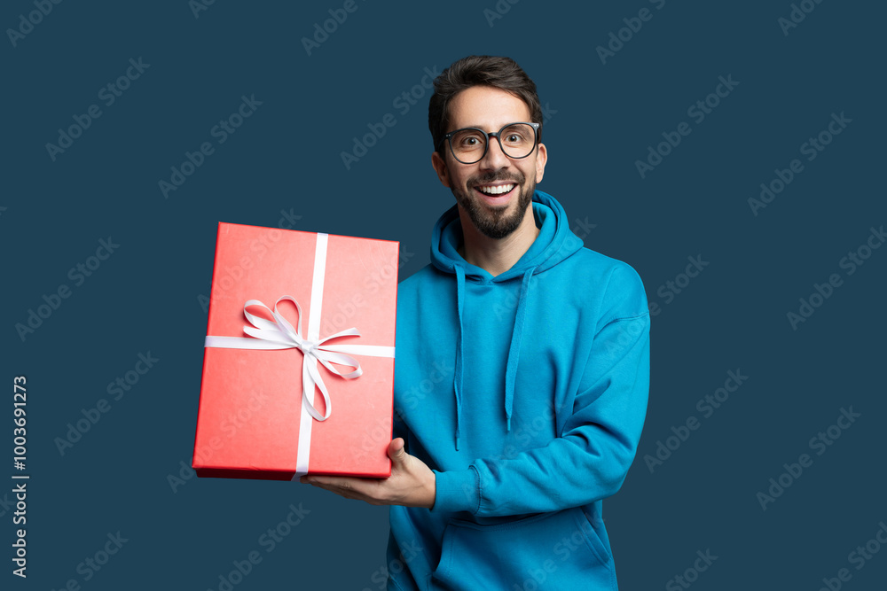 A young man wearing glasses and a blue hoodie happily presents a large red gift box tied with a white ribbon against a solid blue background, displaying excitement and joy. A young man wearing glasses and a blue hoodie happily presents a large red gift box tied with a white ribbon against a solid blue background, displaying excitement and joy.