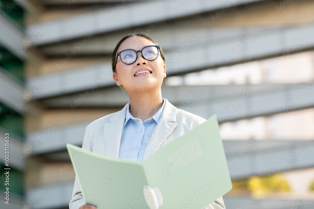 Asian woman dressed in business attire looks up with a smile while holding a folder outside a contemporary building. She appears confident and engaged, enjoying her surroundings. Asian woman dressed in business attire looks up with a smile while holding a folder outside a contemporary building. She appears confident and engaged, enjoying her surroundings.