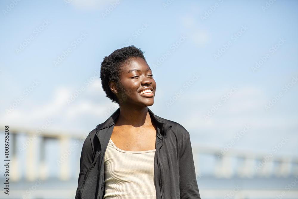 A young African American woman stands by the water under a clear blue sky, smiling joyfully. Behind her, a long bridge stretches across the horizon, creating a picturesque view on a beautiful day. A young African American woman stands by the water under a clear blue sky, smiling joyfully. Behind her, a long bridge stretches across the horizon, creating a picturesque view on a beautiful day.