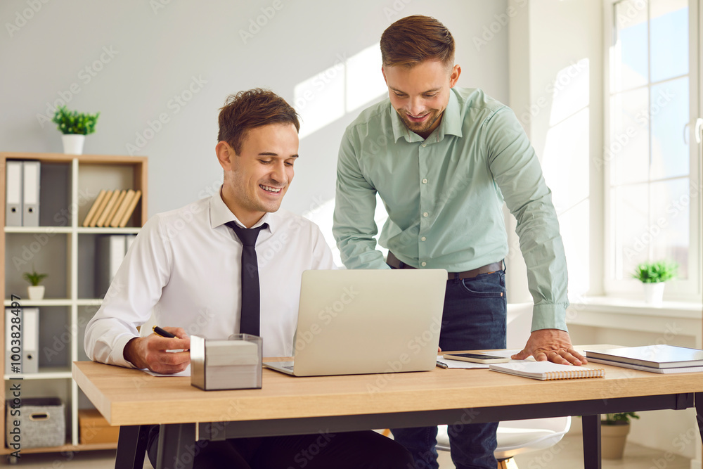 Two men work together. Friendly man gives job consultation to his new colleague in modern office. One man is sitting in front of laptop and other is standing next to him and showing on screen. Two men work together. Friendly man gives job consultation to his new colleague in modern office. One man is sitting in front of laptop and other is standing next to him and showing on screen.