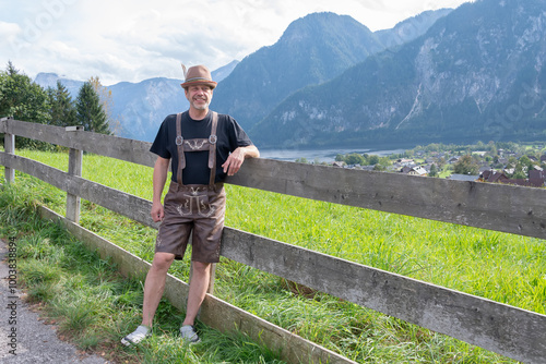 A man in traditional Tyrolean clothing stands by a wooden fence against a backdrop of mountainous landscape and countryside.