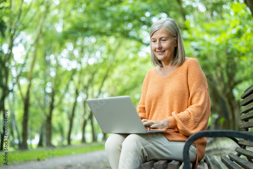 Senior woman sitting on park bench using laptop, smiling as she enjoys nature. Represents outdoor leisure, online work, and digital lifestyle for older adults.