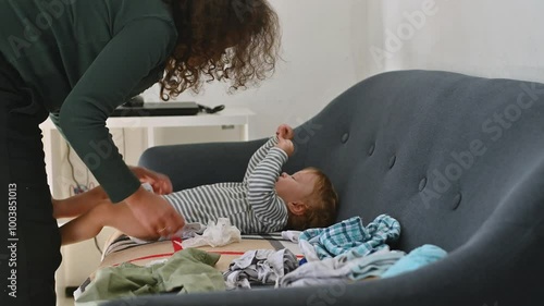 Mother changing baby's diaper on sofa with clothes scattered around in a home setting