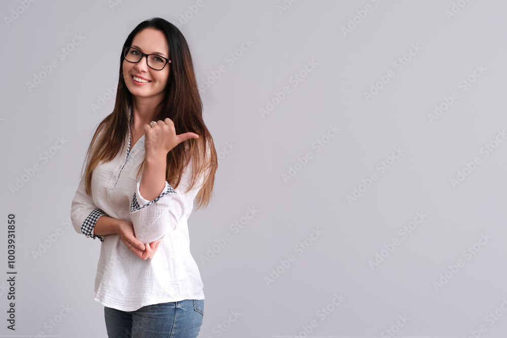 A smiling white woman with long brown hair, wearing glasses and a casual white shirt, pointing to her left with her thumb. She stands against a plain background with copy space A smiling white woman with long brown hair, wearing glasses and a casual white shirt, pointing to her left with her thumb. She stands against a plain background with copy space