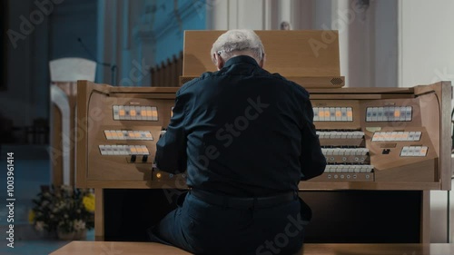 Back view of senior organist sitting at organ in black clothes and playing keyboard instrument