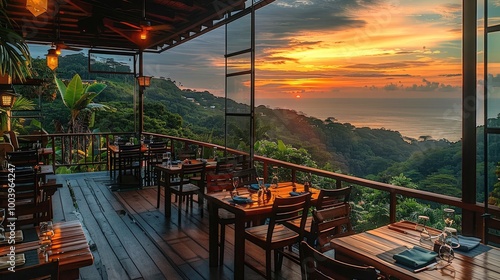 Fototapeta Naklejka Na Ścianę i Meble -  The restaurant is located on the top floor of an apartment building, overlooking the sunset view over jungle-covered hills and the sea in the background. dining tables have wooden chairs around them.