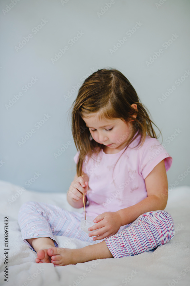 A girl child eats ice cream sitting on the bed. Rejoices, grimaces, enjoys. Holiday, weekend, home A girl child eats ice cream sitting on the bed. Rejoices, grimaces, enjoys. Holiday, weekend, home
