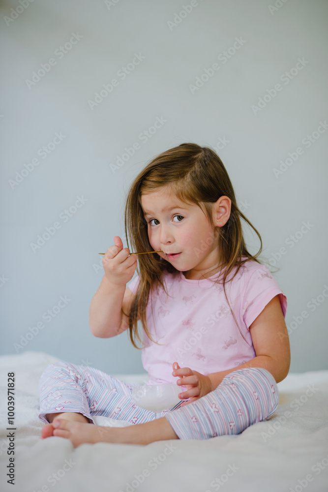A girl child eats ice cream sitting on the bed. Rejoices, grimaces, enjoys. Holiday, weekend, home A girl child eats ice cream sitting on the bed. Rejoices, grimaces, enjoys. Holiday, weekend, home