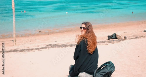 Caucasian Woman in Glasses Sitting on a Sandy Beach, Looking Directly at the Camera. Clear Blue Water On The Background. Work-Life Balance, Nomadic lifestyle photo