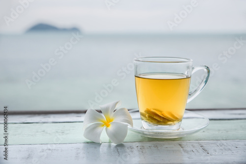 Cup of green tea with ginger servered with plumeria frangipani flower front of at sunset light on wooden tabletop. Sea view.